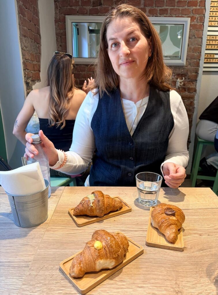 Woman sitting at a cafe table with three gluten-free filled croissants on wooden serving boards, illustrating a functional nutritionist’s guide to a gluten-free lifestyle.