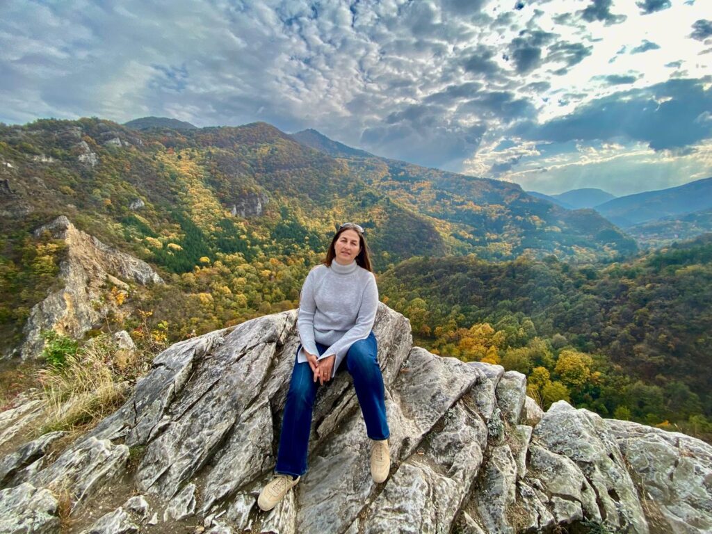A woman sitting on a rugged mountain rock overlooking a vast autumn landscape, symbolising resilience during the journey of menopause, grief, and autoimmunity.